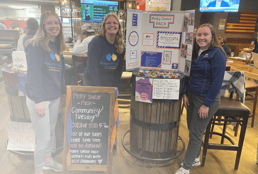 Three college students wearing Pinky Swear shirts raising money for kids with cancer with signs.