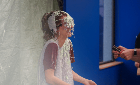 A person sitting in a chair covered in silly string wearing goggles during a Cancer is Messy fundraiser for kids with cancer